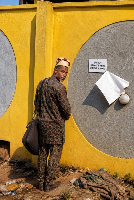 A man in traditional dark patterned attire and a small hat stands against a bright yellow wall with circular designs, near a sign that reads 'DO NOT URINATE HERE FINE IS N20000'.