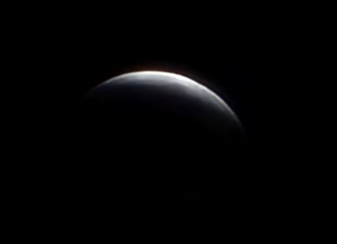 A dark, moody image showing the curved edge of a planet illuminated by a thin sliver of light against a vast black background, resembling a crescent view of Earth from space.