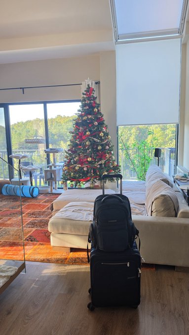 A black backpack sits atop a rolling suitcase in the entryway of a modern home decorated for Christmas with a fully adorned tree, ready for a spontaneous trip. The image captures the excitement of an unplanned getaway, echoing the spontaneity of the movie quote as the traveler prepares to "see about a girl."