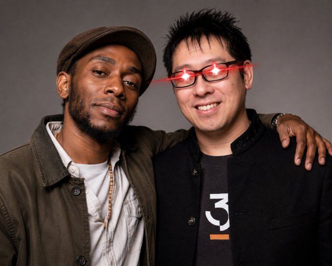 Two men pose closely together for a portrait; the man on the left wears a cap and a green jacket over a white shirt, while the smiling man on the right, wearing glasses that catch the camera flash with red reflections, sports a black jacket over a t-shirt displaying the text "3". The image aligns with the post's "Samson Mos-def" caption, likely depicting Mos Def (Yasiin Bey) on the left in a relaxed, friendly pose with a companion.