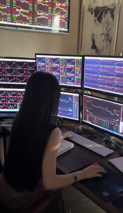 A woman with long black hair sitting at a desk in front of multiple computer monitors displaying various colorful stock market charts and graphs.