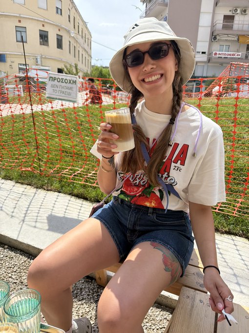 A smiling woman in a bucket hat, sunglasses, and a t-shirt sits on a wooden bench holding an iced coffee, with a 'ΠΡΟΣΟΧΗ ΔΕΝ ΠΥΤΑΜΕ ΤΟ ΓΚΑΖΙ' sign on an orange fence behind her.
