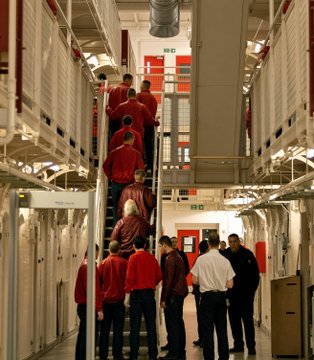 A group of people, some wearing red jackets, are gathered in a narrow corridor between rows of bunk beds in a prison facility. The scene visually illustrates the article's context of prison overcrowding, showing confined living conditions with multiple bunk beds stacked closely together.