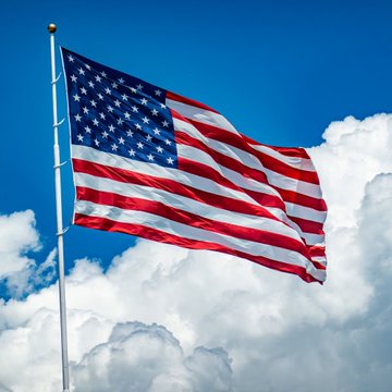 An American flag waves against a blue sky with white clouds, symbolizing the United States as the backdrop for the news that US spot Bitcoin ETFs experienced $57M in net outflows on August 14.