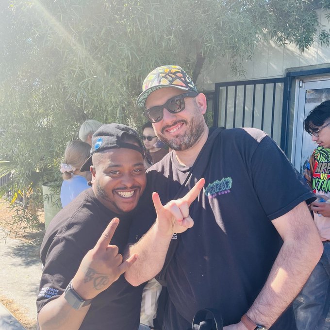 Two smiling men posing together outdoors, with the man on the left wearing a black t-shirt and backwards cap, and the man on the right wearing sunglasses, a cap, and a black t-shirt.