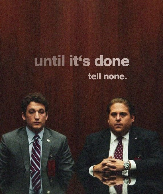 Meme image showing two men in suits sitting at a table against a dark red wall, with text overlaid reading 'until it's done tell none.'