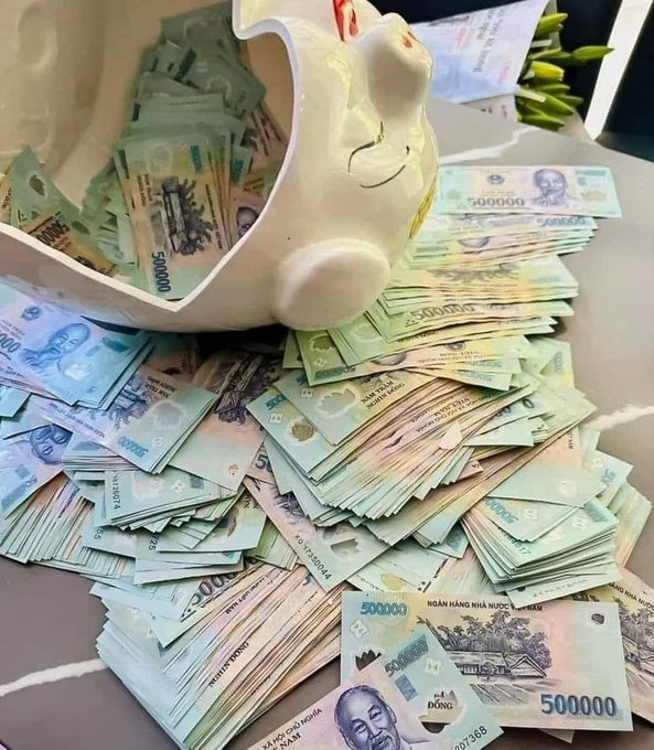 A piggy bank overflowing with stacks of Vietnamese Dong banknotes (500,000 VND denominations) scattered around it on a table.