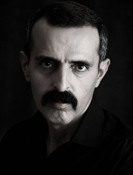 Stark black and white portrait photograph of a man with a thick mustache and short dark hair, gazing intensely at the camera.