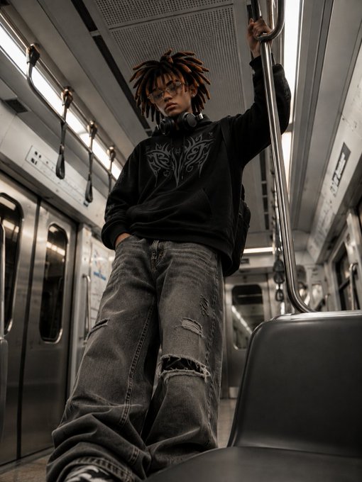A young man with dreadlocks, glasses, and headphones around his neck stands inside a subway car, holding a metal handrail while wearing a black graphic hoodie and distressed baggy jeans.