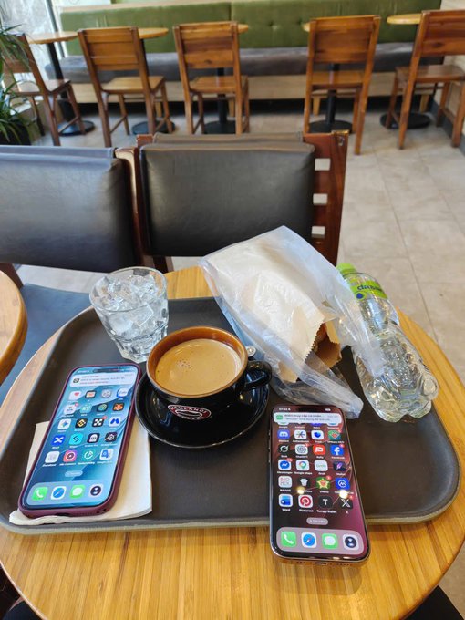 A coffee shop tray holds a black espresso cup on a saucer, a glass of water, a plastic bottle, and two iPhones displaying home screens, all resting on a wooden table with chairs and a booth in the background.