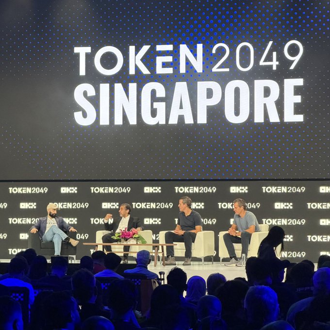 Four panelists discuss cryptocurrency topics on stage at TOKEN2049 Singapore 2024, with the event's logo prominently displayed on a large screen behind them, while an audience watches the conversation unfold.