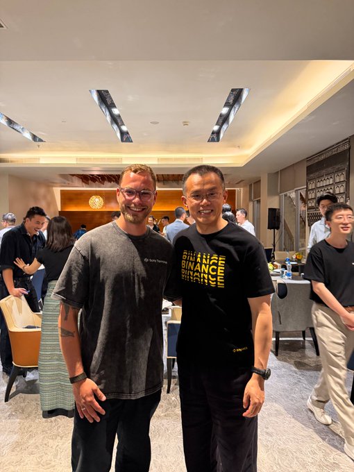 Two men pose together at an indoor event, one wearing a Binance t-shirt with "BINANCE BLOCKCHAIN WEEK" text, while the other wears a gray "Sanaa Hafrabachi" shirt.