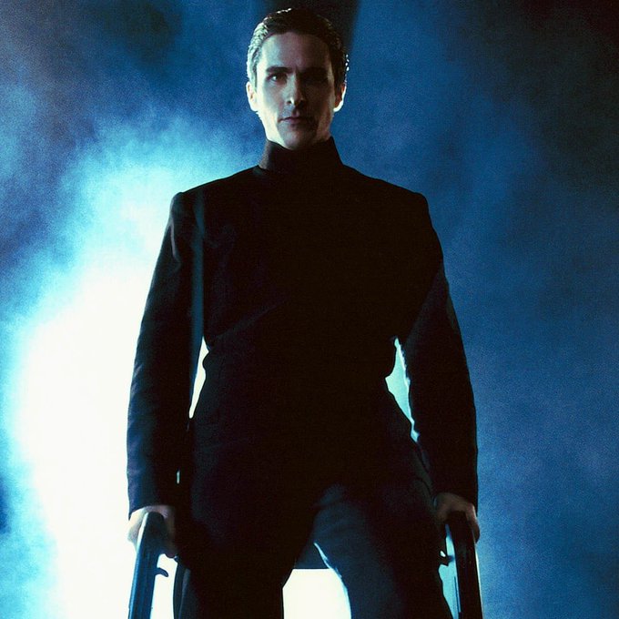 A man in a dark, high-collared suit stands amidst blue haze, gripping a weapon in each hand with an intense, commanding gaze. The image captures the iconic scene from the film *Equilibrium*, where the character Father represents the cold, calculated demeanor that the post playfully references as the perfect moment for an intellectual connection.