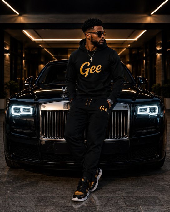 A man wearing sunglasses, a black tracksuit with 'Gee' written in gold script on the hoodie and pants, a gold cross necklace, and black and gold sneakers stands with his arms crossed in front of a black Rolls-Royce.