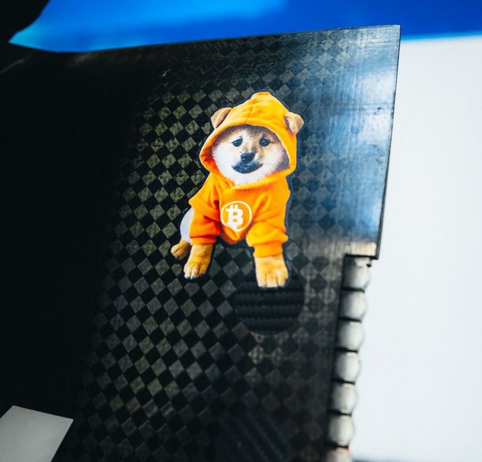 A small figurine or sticker of a Shiba Inu dog wearing an orange Bitcoin (₿) hoodie, placed on a textured black carbon-fiber-like surface with a blue glow in the background.