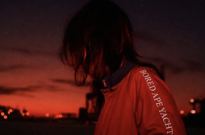 A figure with long hair stands against a deep red twilight sky, wearing a red jacket featuring the white vertical text "BORED APE YACHT" on the sleeve, reflecting the BAYC (Bored Ape Yacht Club) style aesthetic. The dark, moody silhouette and crimson tones evoke the urban, underground vibe associated with the NFT collection.