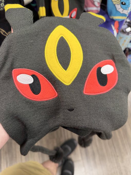 A person holds up a dark gray knit beanie featuring a Pokémon design with large red eyes and a yellow marking on the forehead.
