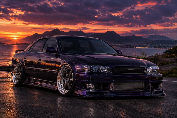 A dark purple modified sedan, appearing to be a Toyota Chaser, parked on a wet surface with mountains and a sunset in the background.