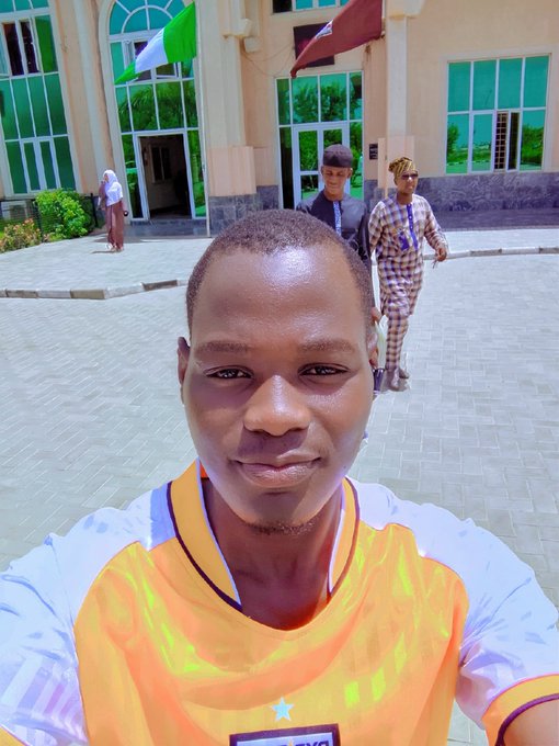 A young man takes a selfie in the foreground wearing a vibrant orange and white soccer jersey with a star emblem and "Bybit" branding, while two men walk in the background near a building displaying flags. The image captures the community aspect mentioned in the post, showing Bybit being represented outdoors in a casual, real-world setting.