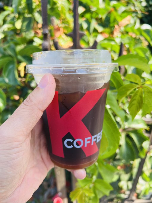 The image features a person holding up a coffee cup with "K" written on it, which appears to be filled with a dark liquid. The background of the photo shows green leaves from trees, suggesting that the setting could be an outdoor area or a park. There is also text in the image related to Twitter and projects, possibly indicating that this person has been sharing their thoughts about these topics online.