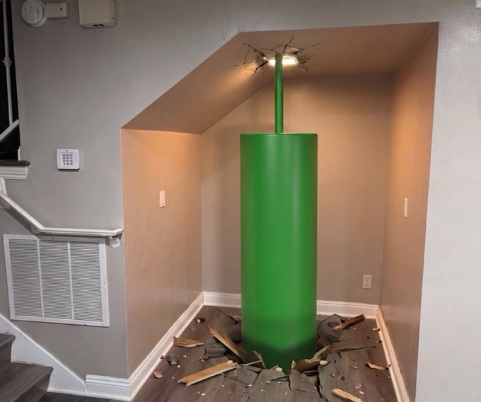 An interior under-stairs nook featuring a green pipe emerging from a hole in the floor and ceiling, styled like the warp pipes from the Super Mario video game series.