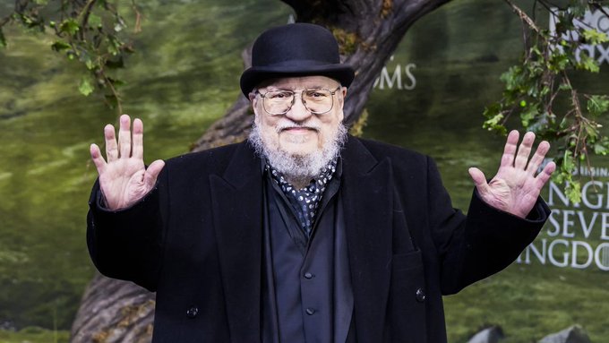 George R.R. Martin, the 'Game of Thrones' author, wearing his signature black bowler hat, glasses, and a dark suit with a patterned cravat, posing with both hands raised in a shrug at what appears to be a 'House of the Dragon' premiere event.