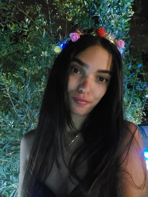 A young woman with long dark hair wears a colorful flower crown adorned with small lights, posing against a lush green leafy background at night, capturing a serene and reflective mood that complements the post's message of gratitude and anticipation for the new week.