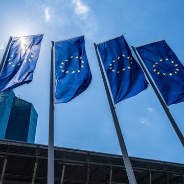 Four European Union flags wave against a bright blue sky in front of what appears to be a modern institutional building, likely symbolizing the EU regulatory framework under which ESMA operates. The flags visually represent the European Union authority behind the MiCA regulations referenced in the post about Germany's Crypto-Asset Service Providers (CASPs).