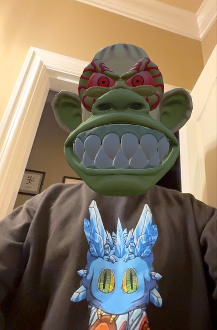 A person wearing a green monkey-like mask and a dark t-shirt featuring a blue ice dragon character, standing indoors.
