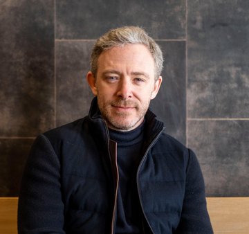 A man with short gray hair and a beard, wearing a dark navy zip-up jacket over a black turtleneck, poses for a portrait against a textured dark stone wall. The image accompanies a post from @dominic_w discussing the ongoing development of advanced smart contracts as a better backend tech stack since 2015, with the ticker $ICP shown.