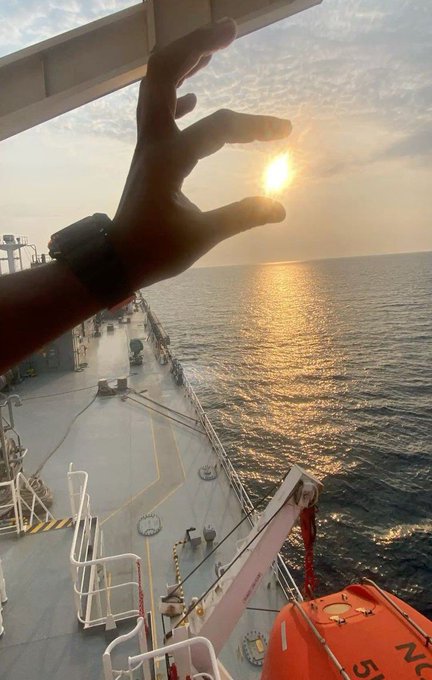 A view from the deck of a ship at sunset, with a hand raised in the foreground, one hand wearing a watch, and a life preserver visible on the right.