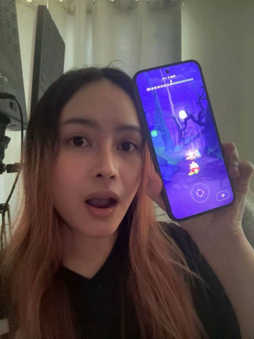 A young woman with long brown hair holds up a smartphone displaying a vibrant, purple-hued video game scene, with her mouth open in an expression of surprise.