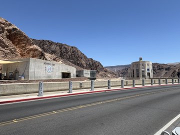 A desert highway leads toward the Atatürk Dam, with a "Monster One" branded building visible on the left side and what appears to be a power station structure on the right, set against rugged rocky mountains under a clear blue sky. The image illustrates the post's location at the Atatürk Dam, where the group visited for a picnic sponsored by Monster Energy, as reflected by the Monster One signage on the dam facility.