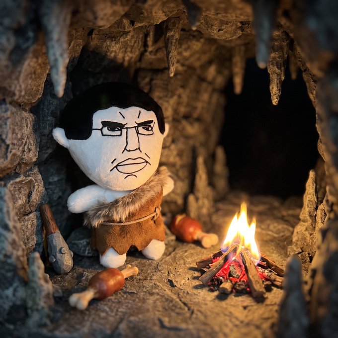 A plush doll with an angry human-like face wearing a fur vest stands in a cave setting next to a small campfire and roasted chicken drumsticks.