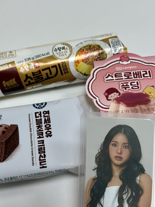 An assortment of Korean snacks including a 'Pent of' B'PROOF brownie and Peripera strawberry pudding pouch are arranged next to a photocard of a woman with long dark hair.