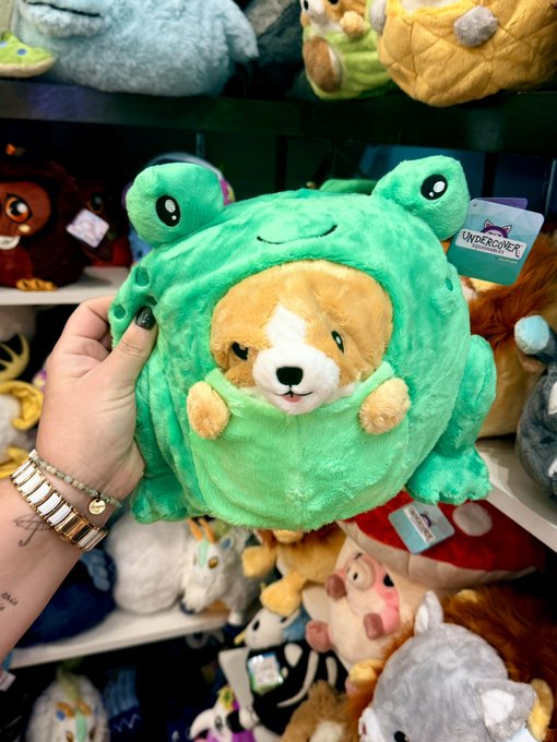 A hand holds a plush dog dressed in a green frog costume in front of shelves of stuffed animals; a tag reads 'UNDERCOVER'.