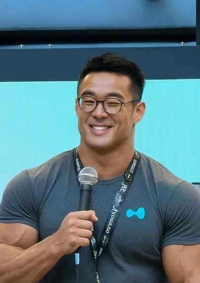 A smiling Asian man with glasses, wearing a grey t-shirt and a lanyard, holding a microphone while speaking at an event with a teal background.