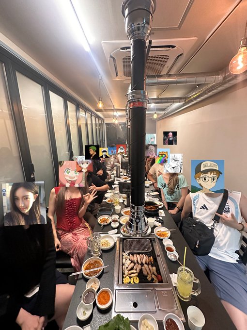 A lively group of friends gather around a table at a Korean BBQ restaurant, featuring a central exhaust pipe, grilled meat on the grill, and an array of side dishes and sauces. The image ties to a celebratory dinner gathering, with attendees' faces playfully covered by cartoon and celebrity images, capturing the social spirit ahead of the upcoming MemeCore Lotte World event.