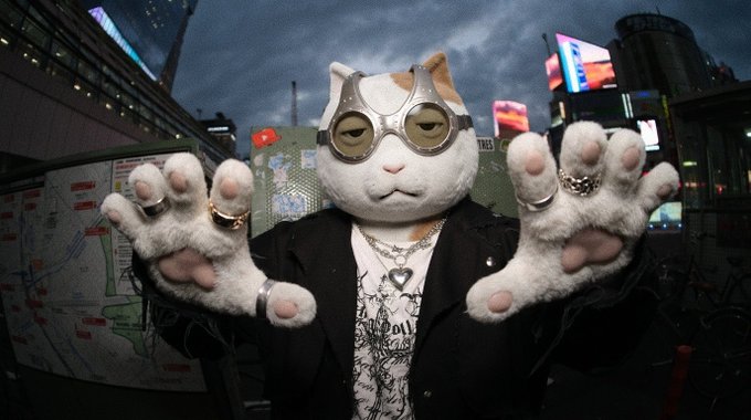 Costumed cat character wearing goggles, a black jacket, and multiple rings and necklaces, holding both paws up toward the camera against an urban backdrop.