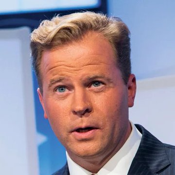 A portrait shows a blonde-haired man with blue eyes wearing a dark suit and tie, captured mid-speech against a blurred background.