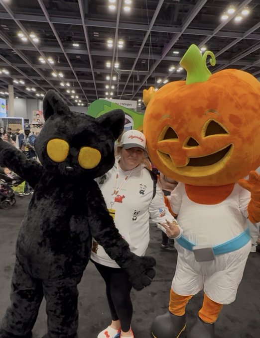 Photo of a woman in a white cap and shirt posing between two mascots—a black cat with large yellow eyes and an orange jack-o'-lantern character—at what appears to be a convention.