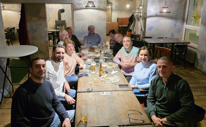 A group of eight people sitting around a long wooden table in a restaurant with industrial-style decor, posing for a photo with food and drinks on the table.