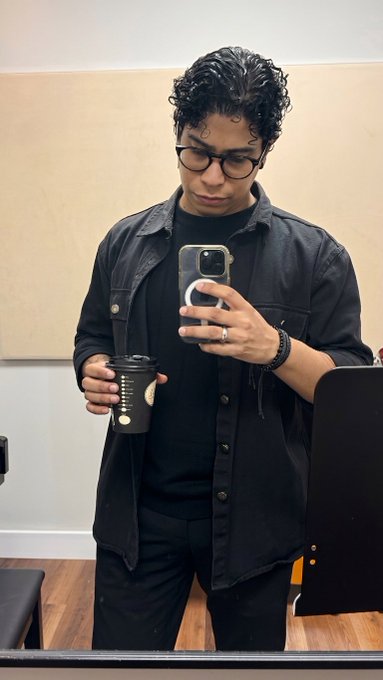 A mirror selfie of a young man with curly dark hair and glasses, wearing a black button-up shirt and holding a coffee cup, with a PopSocket on his phone.