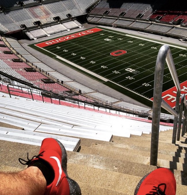 A first-person perspective shot showing a person wearing red Nike sneakers sitting on concrete steps high up in an empty Ohio State Buckeyes stadium, looking down at the football field.