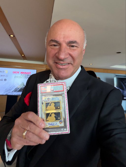 Kevin O'Leary, known as "Mr. Wonderful," smiles while holding up a collectible gold-themed card display at the Avalanche Summit NYC. The event is advertised in the background screen, where he is scheduled to speak live at 12:05pm.