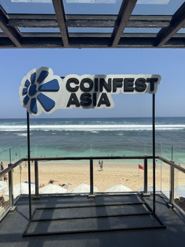A sign for 'COINFEST ASIA' with a blue flower-like logo stands on a beachfront patio overlooking the ocean.