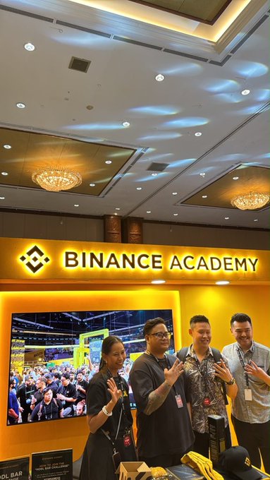 A photo of four people posing in front of a yellow 'BINANCE ACADEMY' booth with an illuminated logo at what appears to be a conference.