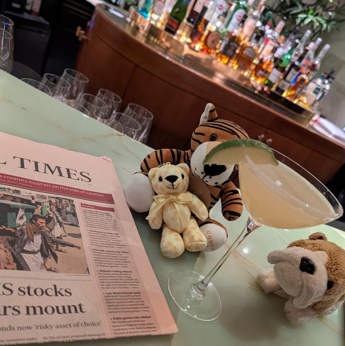 A newspaper titled 'TIMES' with headlines about 'stocks' and 'investors mount' lies next to a cocktail garnished with lime, accompanied by three stuffed animals including a tiger and a bear, set in front of a bar.