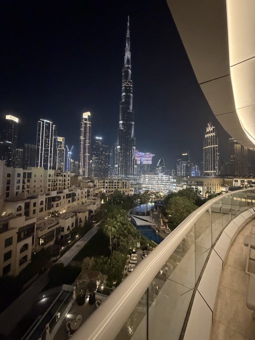 Nighttime view of the Burj Khalifa and Dubai skyline from a curved balcony, with illuminated skyscrapers and a pool area below.