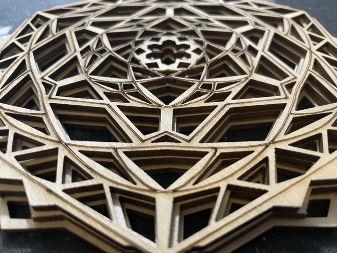 An intricate laser-cut wooden mandala or geometric design featuring multiple layers of triangular and hexagonal patterns arranged in a circular mandala formation.